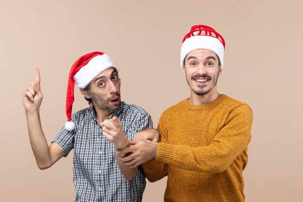 Vorderansicht zwei lächelnde Kerle mit Weihnachtsmützen, von denen einer etwas zeigt, das andere seine Hand auf beige lokalisiertem Hintergrund hält