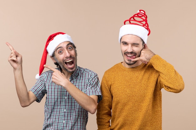 Vorderansicht zwei lächelnde Kerle mit Weihnachtsmützen, von denen einer etwas zeigt und der andere mich Telefonschild auf beigem isoliertem Hintergrund anruft