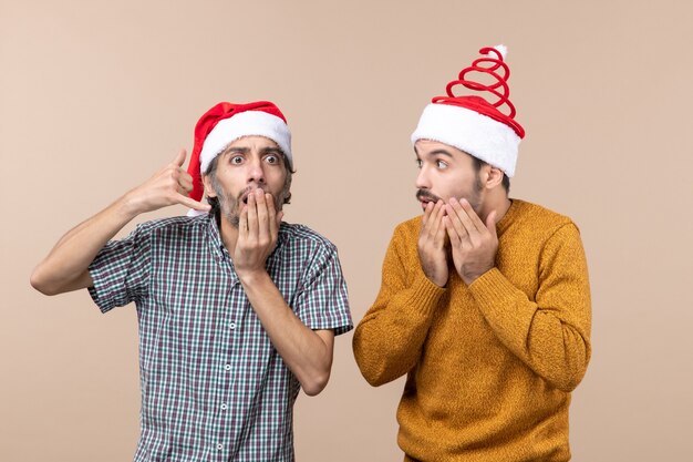 Vorderansicht zwei seltsame Kerle mit großer Überraschung, die Weihnachtsmützen auf beigem isoliertem Hintergrund tragen