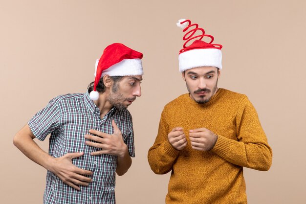 Vorderansicht zwei verwirrte Kerle mit Weihnachtsmützen, die einen die andere Hand auf beigem isoliertem Hintergrund betrachten