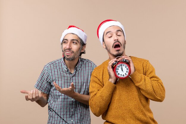 Vorderansicht zwei Weihnachtsmänner, die einen Wecker mit geschlossenen Augen auf beige lokalisiertem Hintergrund halten