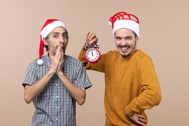 Vorderansicht zwei Weihnachtsmänner eine überraschte und eine glückliche, die einen Wecker auf beige lokalisiertem Hintergrund hält