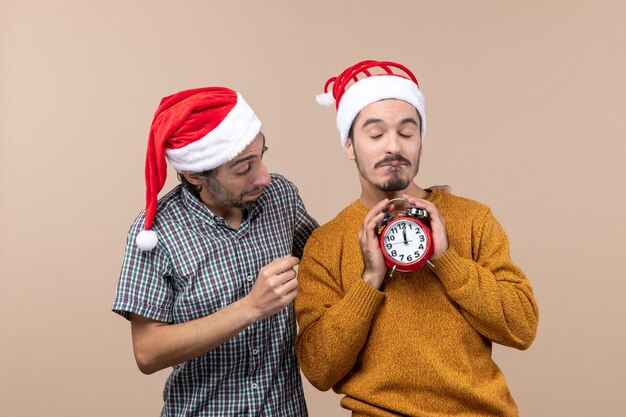 Vorderansicht zwei Weihnachtsmänner eins, die einen Wecker mit beiden Händen auf beige lokalisiertem Hintergrund halten