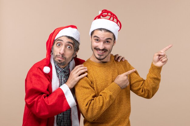 Vorderansicht zwei Weihnachtsmänner mit Weihnachtsmützen, die eine Freundschulter halten und die andere Richtung auf beige lokalisiertem Hintergrund zeigen