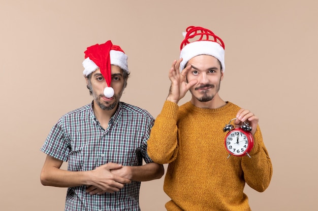 Vorderansicht zwei Weihnachtsmänner, von denen einer Hände an seine Brust legt und der andere einen Wecker auf beigem isoliertem Hintergrund hält