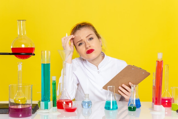 Vordere Ansicht junge Chemikerin im weißen Anzug mit ed-Lösungen, die mit ihnen arbeiten, die Notizblock auf dem gelben Raumchemie-Wissenschaftsprozess halten