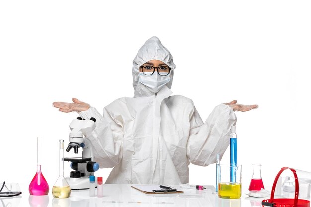 Vordere Ansicht junge Chemikerin in speziellem Schutzanzug sitzend mit Lösungen auf weißem Hintergrundviruslabor Covid Health Chemistry