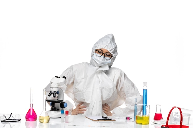 Vordere ansicht junge chemikerin in speziellem schutzanzug sitzt mit tisch voller lösungen auf weißer hintergrundviruslabor-covid-chemie