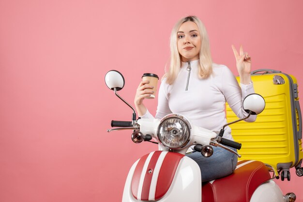 Vordere Ansicht junge Dame auf Moped hält Kaffeetasse, die Siegeszeichen macht