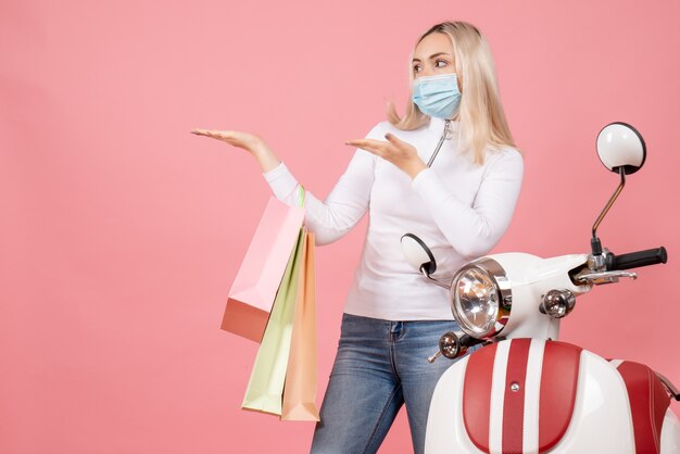 Vordere Ansicht junge Dame, die Einkaufstaschen hält, die links nahe Moped auf rosa Wand zeigen