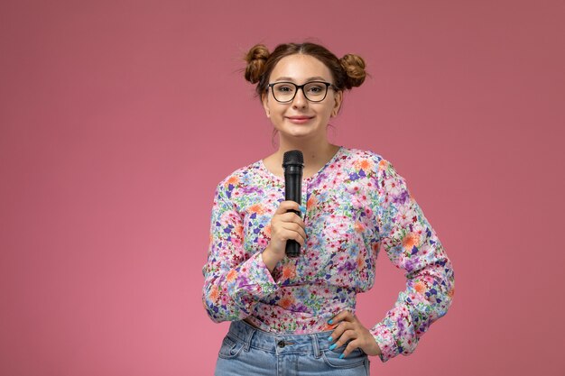 Vordere Ansicht junge Frau in Blume entworfenes Hemd und blaue Jeans, die versuchen, mit Mikrofon auf rosa Hintergrund zu singen