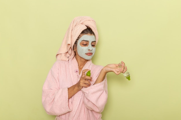 Vordere Ansicht junge Frau nach der Dusche im rosa Bademantel, der Sprays auf grüner Oberfläche hält