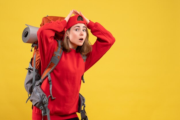 Vordere Ansicht junge Reisende Frau im roten Rucksack, der Hände über ihrem Kopf verbindet, der auf gelber Wand steht