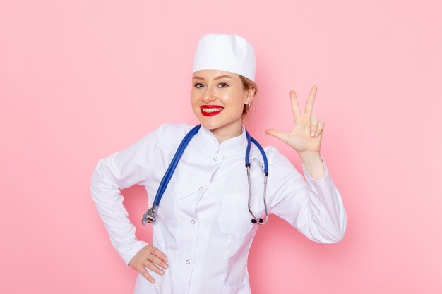 Vordere Ansicht junge Ärztin im weißen Anzug mit blauem Stethoskop lächelnd und posierend auf der Ärztin des rosa Raummedizin-medizinischen Krankenhausarztes