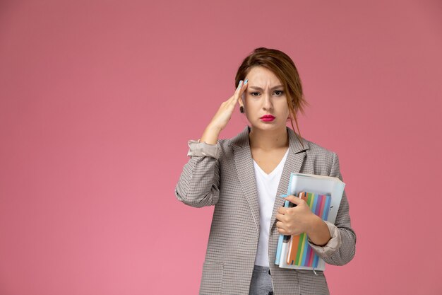 Vordere Ansicht junge Studentin im grauen Mantel, der Hefte aufstellt und hält, die Kopfschmerzen auf rosa Hintergrundstunden Universitätsuniversitätsstudie haben