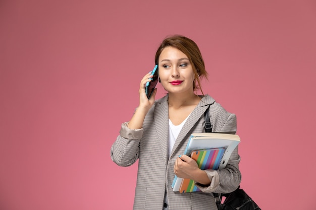 Vordere Ansicht junge Studentin im grauen Mantel mit Heften, die am Telefon auf rosa Hintergrundstunden Universitätsuniversitätsstudie sprechen