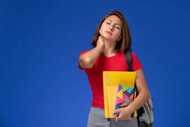 Vordere Ansicht junge Studentin im roten Hemd, das Rucksack hält, der Dateien und Heft hält, das Nackenschmerzen auf blauem Hintergrund hat.
