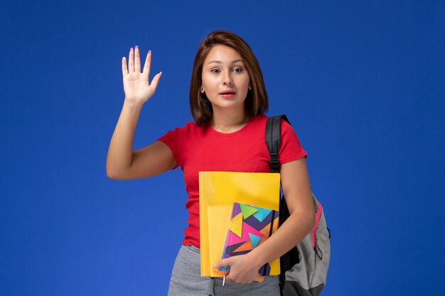 Vordere Ansicht junge Studentin im roten Hemd, das Rucksack hält, der Dateien und Heft hält, die auf blauem Hintergrund winken.