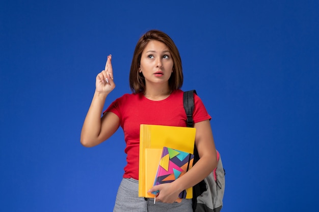 Vordere Ansicht junge Studentin im roten Hemd, das Rucksack hält, der Dateien und Heft hält, die ihre Finger auf blauem Schreibtisch kreuzen.