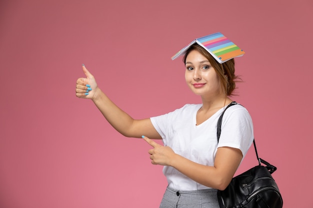 Vordere Ansicht junge Studentin im weißen T-Shirt mit Heft und Tasche, die auf rosa Hintergrundlektionuniversitätshochschulstudienbuch aufwirft und lächelt