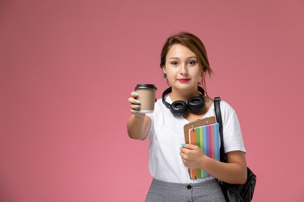 Vordere Ansicht junge Studentin im weißen T-Shirt mit Tasche und Kopfhörern, die Kaffee auf dem rosa Hintergrundlektionsuniversitätsuniversitäts-Studienbuch posieren und lächeln