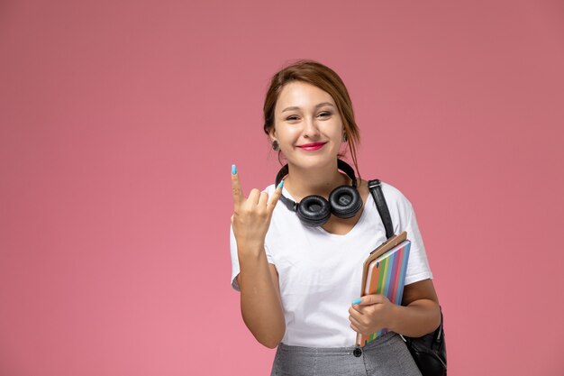 Vordere Ansicht junge Studentin im weißen T-Shirt mit Tasche und Kopfhörern Rocker-Stil posiert und lächelt auf rosa Hintergrund Lektion Universität College-Studienbuch