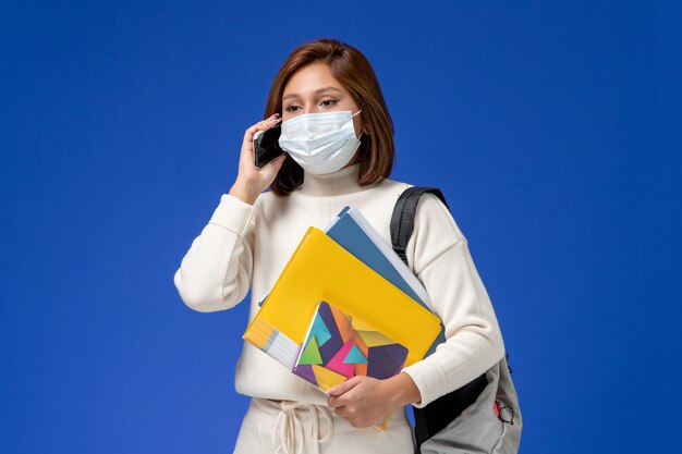 Vordere Ansicht junge Studentin im weißen Trikot mit Maske mit Tasche und Heften, die am Telefon auf blauer Wand sprechen