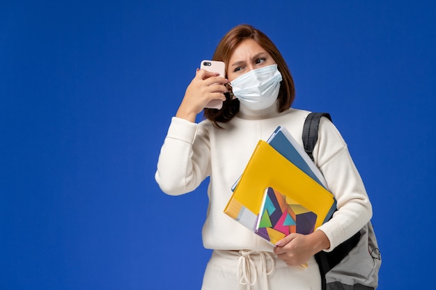Vordere Ansicht junge Studentin im weißen Trikot mit Maske mit Tasche und Heften, die am Telefon auf blauer Wand sprechen