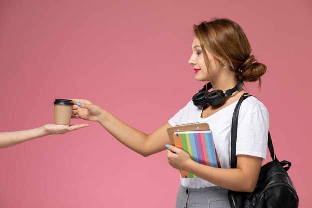 Vordere Ansicht junge Studentin in weißem T-Shirt mit Heft und Tasche, die Kaffeetasse auf dem rosa Hintergrundlektionsuniversitätsuniversitäts-Studienbuch aufwirft und nimmt