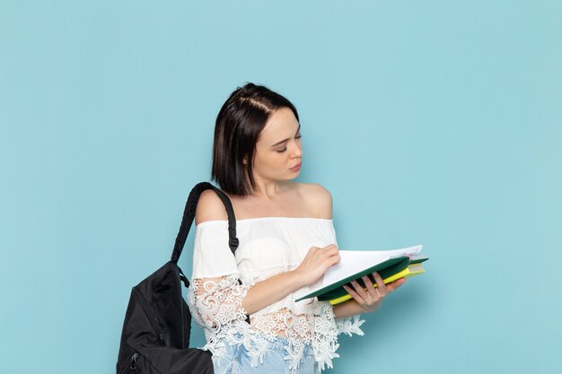 Vordere Ansicht junge Studentin in weißen Hemd Blue Jeans und schwarze Tasche Lesekopierbuch auf dem blauen Raum weibliche Studentin