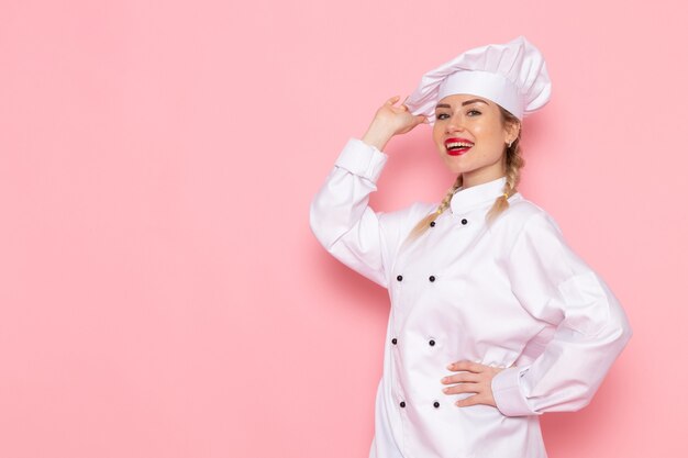 Vordere Ansicht junge weibliche Köchin im weißen Kochanzug, der gerade mit Lächeln auf dem rosa Raumkochküchenjobarbeitsfoto aufwirft