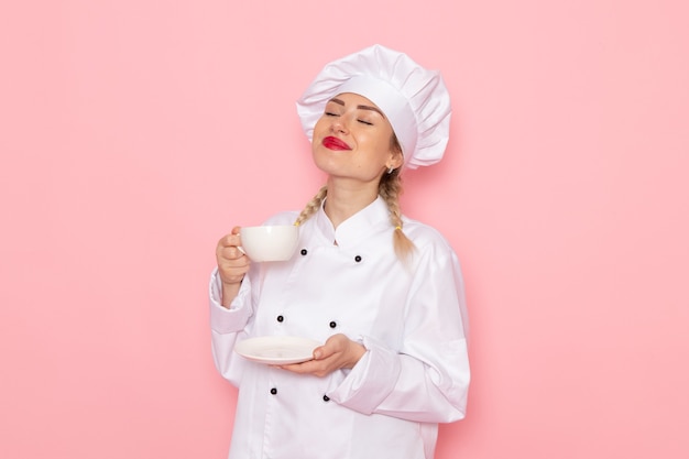 Vordere Ansicht junge weibliche Köchin im weißen Kochanzug, der Tasse Tee auf dem rosa Raumkochfoto hält