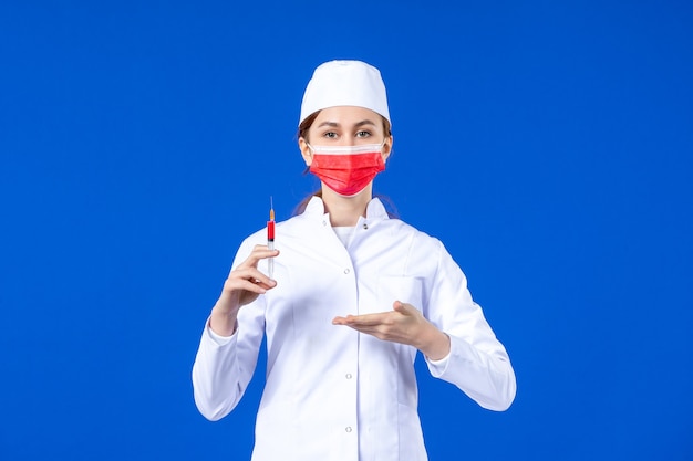 Vordere Ansicht weibliche Krankenschwester im weißen medizinischen Anzug mit roter Maske und Injektion in ihren Händen auf blau