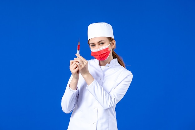 Vordere Ansicht weibliche Krankenschwester im weißen medizinischen Anzug mit roter Maske und Injektion in ihren Händen auf blau