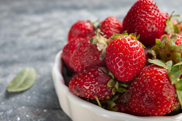 Vordere nähere Ansicht frische rote Erdbeeren innerhalb weißer Platte zusammen mit grünen getrockneten Blättern auf grauem Schreibtisch