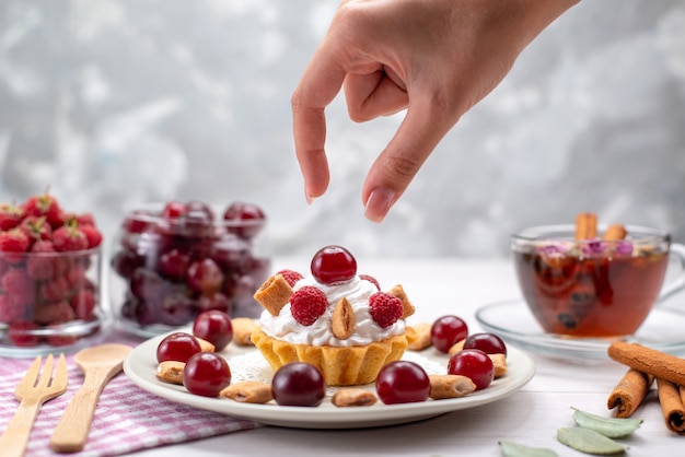 Vordere nähere Ansicht kleiner cremiger Kuchen mit Himbeerkirschen und kleinen Keksen Teezimt auf weißem Schreibtisch