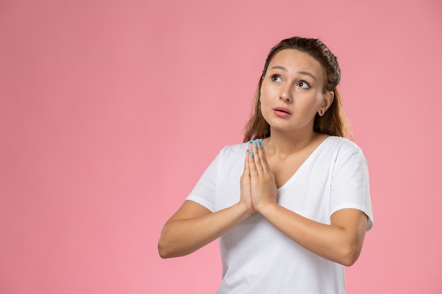 Vordere Nahansicht junge attraktive Frau im weißen T-Shirt mit Gebetshaltung auf dem rosa Hintergrund