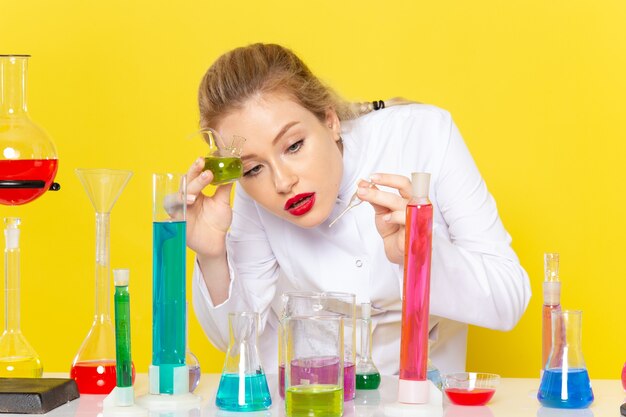 Vordere Nahansicht junge Chemikerin im weißen Anzug vor dem Tisch mit farbigen Lösungen auf gelb