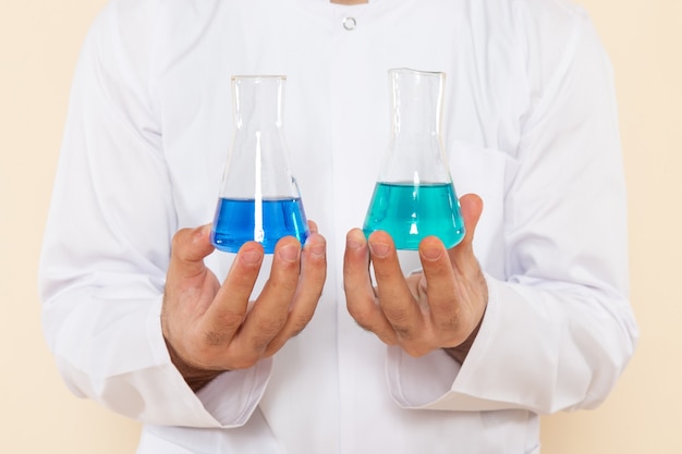 Vordere Nahansicht junger männlicher Chemiker im weißen Spezialanzug, der kleine Flaschen mit Lösungen auf Cremewandwissenschaftsexperimentchemie wissenschaftlich hält