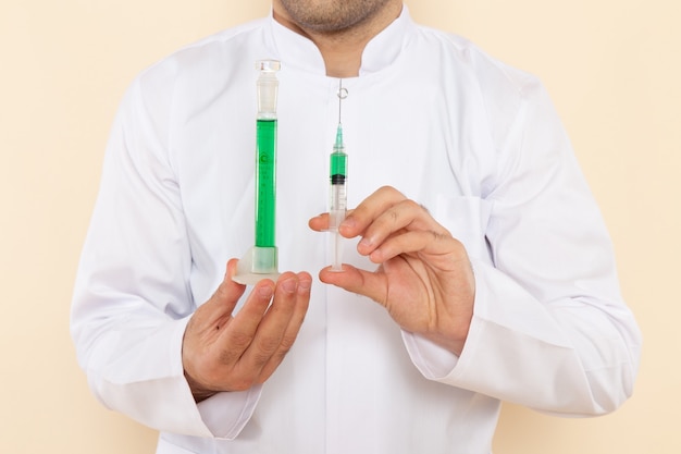 Vordere Nahansicht junger männlicher Chemiker im weißen Spezialanzug, der kleinen Kolben mit grüner Lösung und Injektion auf dem wissenschaftlichen Experimentlabor der Cremewand hält