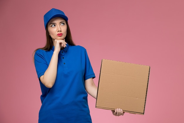 Vorderer Blick junger weiblicher Kurier in der blauen Uniform und im Umhang, der Nahrungsmittellieferbox hält, der an die rosa Wand denkt