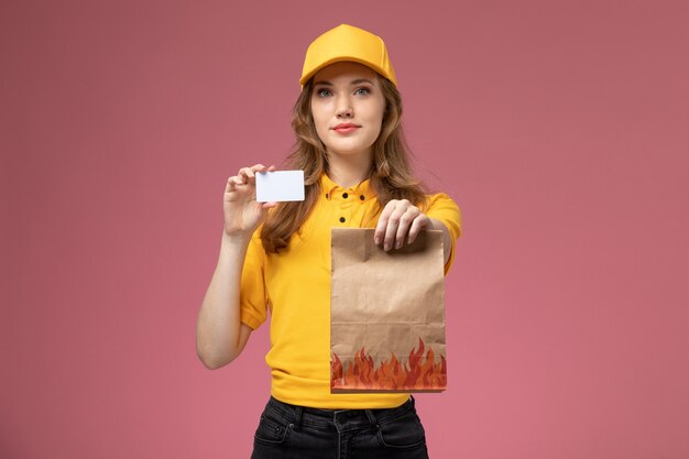 Vorderer Blick junger weiblicher Kurier in der gelben Uniform, die Lieferung Lebensmittelpaket und Plastikkarte auf rosa Hintergrundlieferdienstuniform weiblicher Arbeiter hält