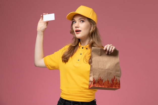 Vorderer Blick junger weiblicher Kurier in gelbem einheitlichem gelbem Umhang, der Lebensmittel-Lieferpaket mit Karte auf der dunkelrosa Schreibtischuniform-Lieferauftragsdienstfarbe hält