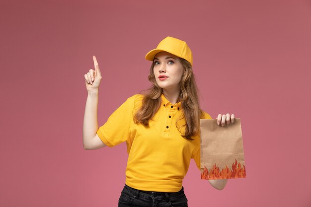 Vorderer Blick junger weiblicher Kurier in gelbem einheitlichem gelbem Umhang, der Nahrungsmittellieferpaket auf dem dunkelrosa Hintergrunduniformlieferdienst weiblicher Arbeiter hält