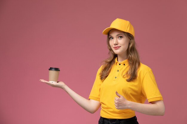 Vorderer Blick junger weiblicher Kurier in gelbem einheitlichem gelbem Umhang, der Plastikkaffeetasse auf der dunkelrosa Hintergrunduniform-Lieferauftragsdienstfarbe hält