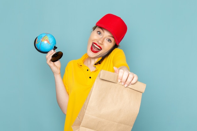 Vorderer Blick junger weiblicher Kurier in gelbem Hemd und rotem Umhang, der Globuspaket hält, das am Telefon auf dem blauen Raum spricht