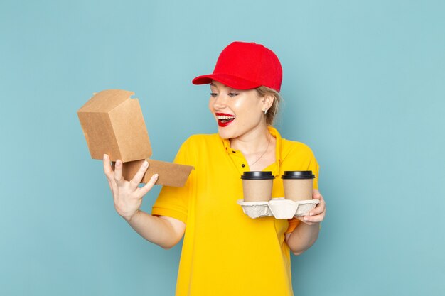 Vorderer Blick junger weiblicher Kurier in gelbem Hemd und rotem Umhang, der Plastikkaffeetassen und Lebensmittelpaket hält, die auf dem blauen Raum lächeln