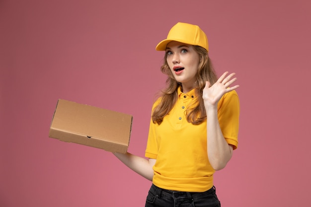 Vorderer Blick junger weiblicher Kurier in gelber Uniform, die Lieferpaket mit überraschtem Ausdruck auf dem rosa Schreibtischjobuniform-Lieferservicearbeiter hält