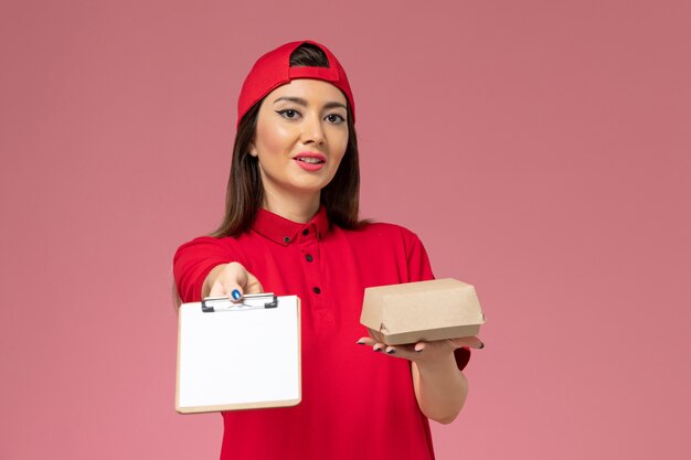Vorderer Blick junger weiblicher Kurier in rotem Uniformumhang mit kleinem Liefernahrungsmittelpaket und Notizblock mit Stift auf ihren Händen an der hellrosa Wand