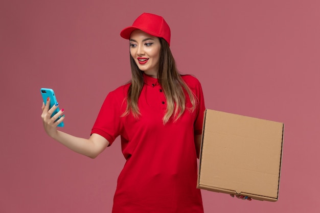 Vorderer Blick junger weiblicher Kurier in roter Uniform, die Lieferung Nahrungsmittelbox und Telefon auf rosa Hintergrunddienstlieferuniform hält